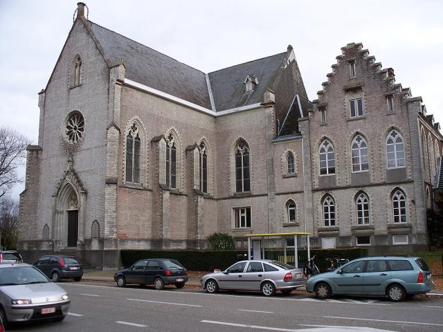 Foto van een groot beige gebouw: het klooster Terbank. Met auto's geparkeerd langs de straat en een bushokje. 